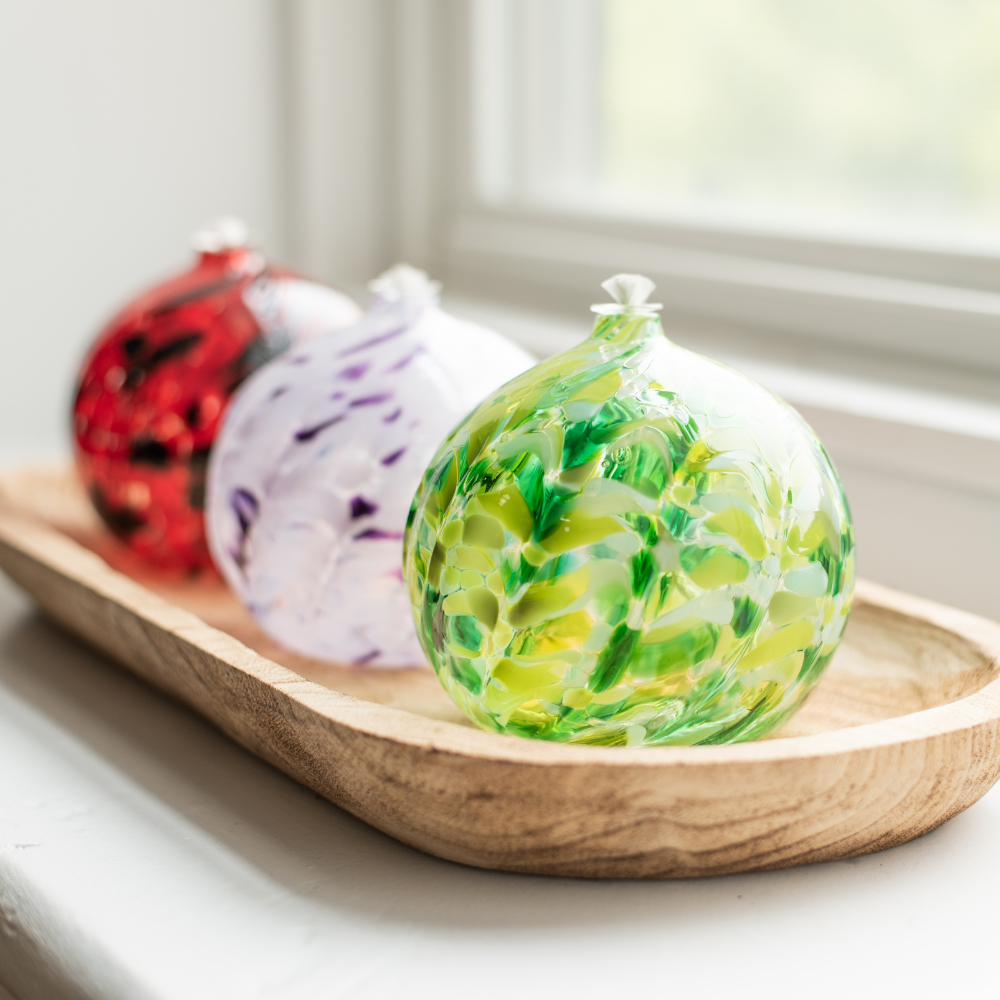 Light and dark green round oil lamp, purple and white round oil lamp and red and back oil lamp on a wood tray on a white window sill