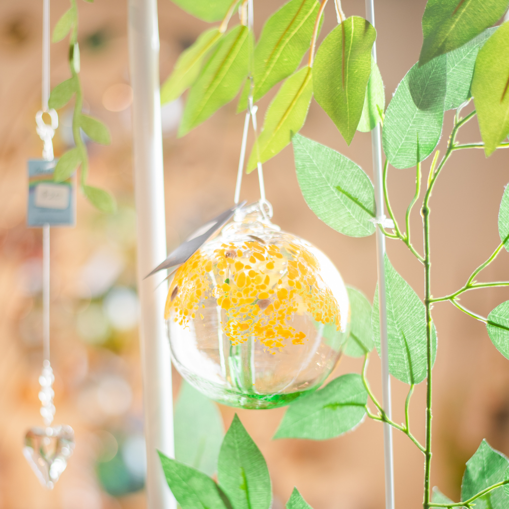 Clear glass orb with a green stem pulled from bottom to top to a canopy of yellow opaque glass blossoms and brown centres hanging amongst greenery. 