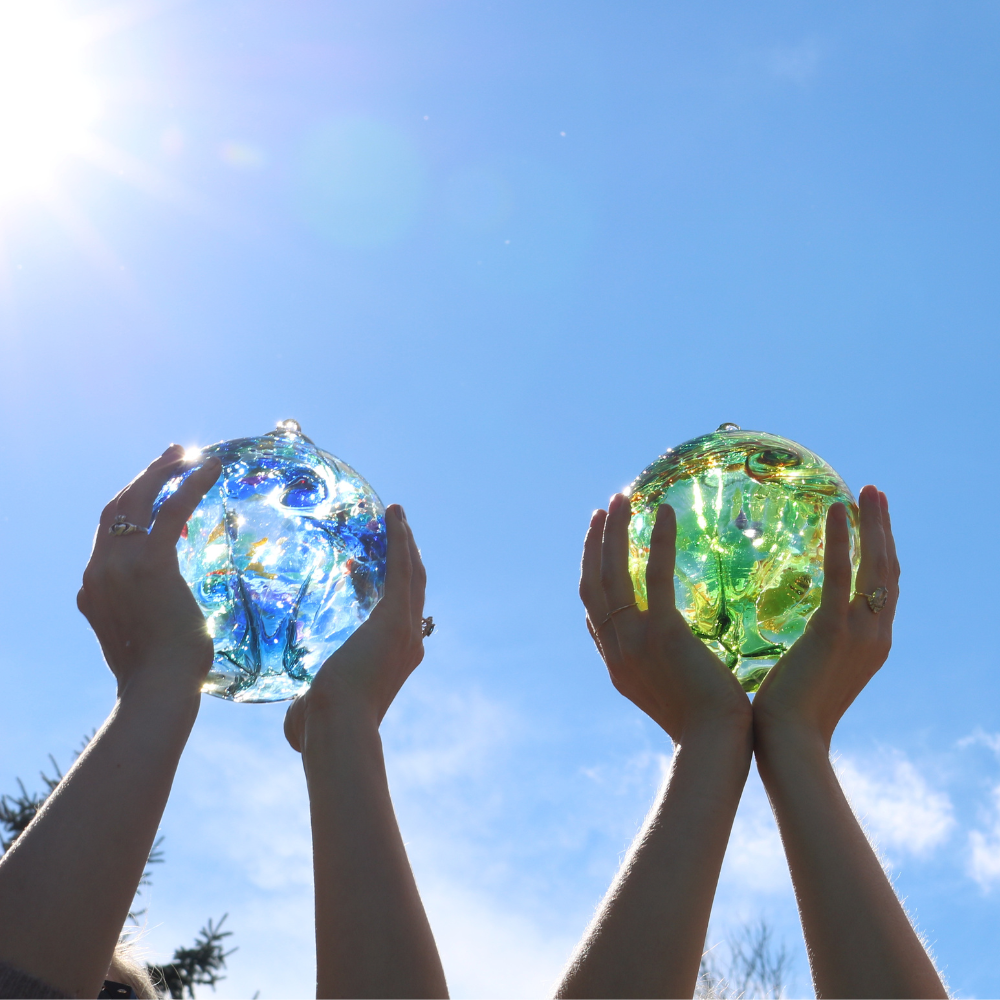 Blue orb and green orb, each held in two hands with a blue sky background and sun shining from the left. 